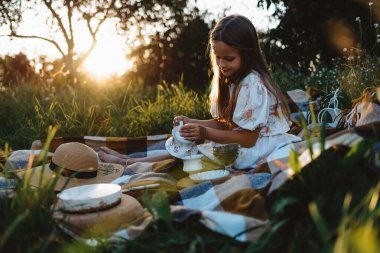 Gün batımında bir kız battaniyeye oturmuş kendine çay dolduruyor.