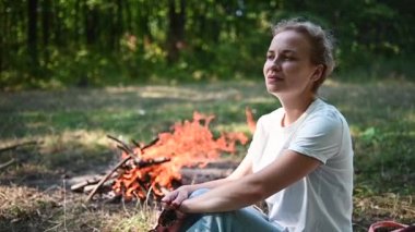 Ormanda ateşin yanında oturan genç bir kadın yukarı bakıyor.