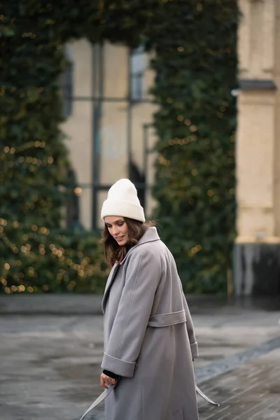 Pretty girl is dancing in gray coat and white hat on the street. Winter outfit. Christmas concept. Place for text