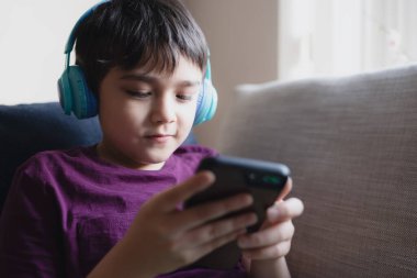 Primary school Kid using mobile phone searching information on internet for homework,Child wearing headphone listening to music,Home schooling,E-learning education,Children with technology