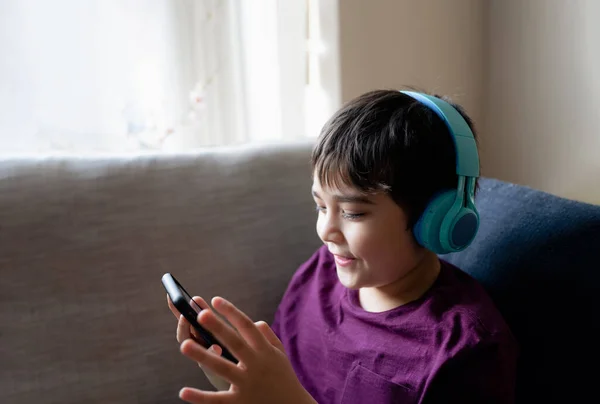 Primary school Kid using mobile phone searching information on internet for homework,Happy Child  boy wearing headphone listening to music,Home schooling,E-learning education,Children with technology
