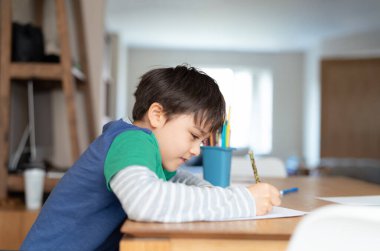 Eğitim, okul öncesi çocuğun beyaz kağıda resim çizmeyi öğrenmesi, kalemle evde deftere yazan mutlu gülümseyen öğrenci, ödev yapan ve sınavlara hazırlanan çocuk..