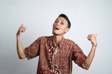 young asian man with brown skin wearing a batik shirt happy expression with joy and showing a thumbs up if he agrees, good, topnotch, white background.