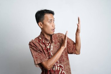 Excited Asian man wearing batik shirt pointing to copy space beside him, isolated on white background