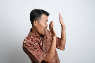Excited Asian man wearing batik shirt pointing to copy space beside him, isolated on white background