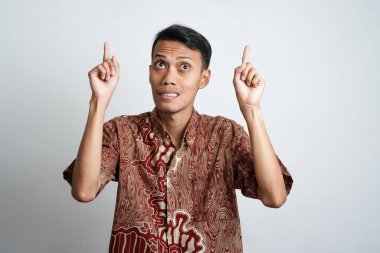 Şok olmuş, batik tişörtlü bir Asyalı beyaz arka planla izole edilmiş, üzerindeki fotokopi alanını işaret ediyor.