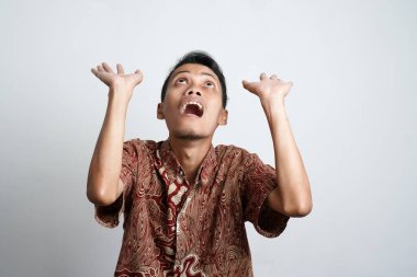 Şok olmuş, batik tişörtlü bir Asyalı beyaz arka planla izole edilmiş, üzerindeki fotokopi alanını işaret ediyor.