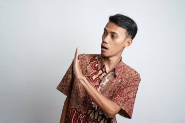 Excited Asian man wearing batik shirt pointing to copy space beside him, isolated on white background