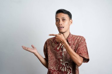 Asian young man with tan skin wearing batik shirt pointing to the side empty space for advertising needs, white background.