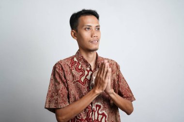 Batik tişört giyen gülümseyen Asyalı genç adam beyaz arka planda izole edilmiş geleneksel selamlaşmalar yapıyor.