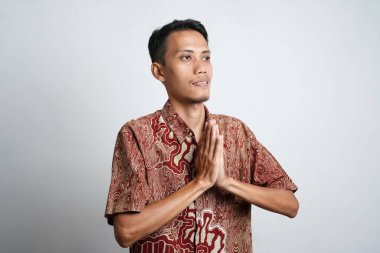 Batik tişört giyen gülümseyen Asyalı genç adam beyaz arka planda izole edilmiş geleneksel selamlaşmalar yapıyor.