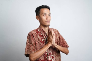 Batik tişört giyen gülümseyen Asyalı genç adam beyaz arka planda izole edilmiş geleneksel selamlaşmalar yapıyor.