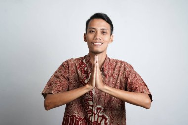 Batik tişört giyen gülümseyen Asyalı genç adam beyaz arka planda izole edilmiş geleneksel selamlaşmalar yapıyor.
