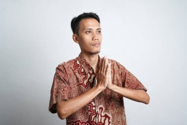 Batik tişört giyen gülümseyen Asyalı genç adam beyaz arka planda izole edilmiş geleneksel selamlaşmalar yapıyor.
