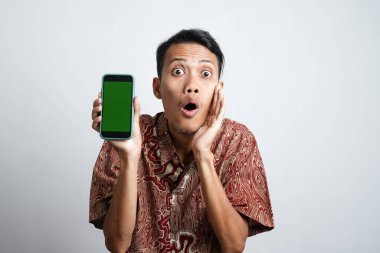 Şok olmuş Asyalı adam batik tişört giyiyor ve telefonunda beyaz arka planla izole edilmiş yeşil ekranı gösteriyor.