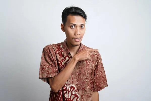 Asian young man with tan skin wearing batik shirt pointing to the side empty space for advertising needs, white background.