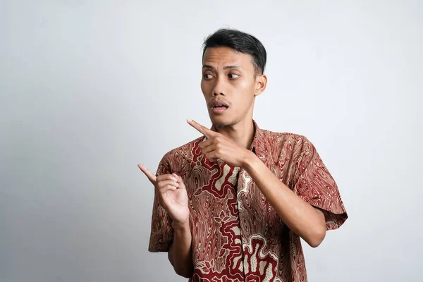 Asian young man with tan skin wearing batik shirt pointing to the side empty space for advertising needs, white background.