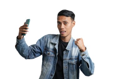 Asian man wearing blue jean jacket doing victory celebration holding smartphone. Isolated by white background