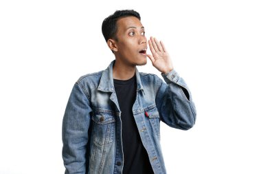 asian man wearing blue jean jacket shouting giving information with hand near mouth, isolated on white background