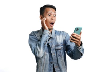 Asian man wearing blue jean jacket surprised expression looking at smartphone screen. Isolated by white background
