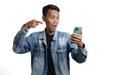 Asian man wearing blue jean jacket surprised expression looking at smartphone screen. Isolated by white background
