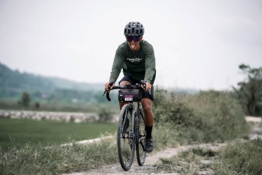 Cyclist in helmet rides bicycle on country road. Using a gravel bike through the rice fields route. : Yogyakarta, Indonesia - 28 January 2023