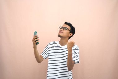 Sıradan çizgili tişört giyen Asyalı bir adamın portresi. Mutlu yüz ifadesi, sürpriz, akıllı telefon tutarken hediye ya da iyi haber almak. Ayrı bej arkaplan.