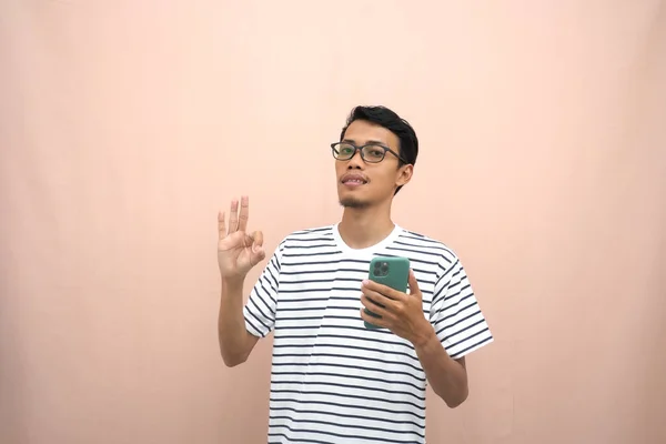 一个亚洲男人戴着眼镜 穿着休闲条纹T恤的肖像 展示大拇指 同意和持有智能手机 孤立的米黄色背景 — 图库照片