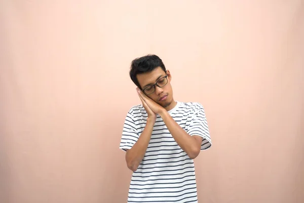 portrait of an asian man wearing glasses wearing a casual striped t-shirt. Sleepy pose, want to sleep, lack of sleep. Isolated beige background.