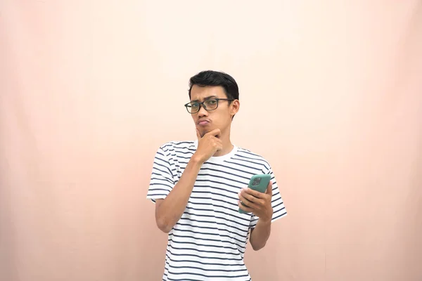 一个亚洲男人戴着眼镜 穿着休闲条纹T恤的肖像 假装在想 想办法拿着智能手机 孤立的米黄色背景 — 图库照片