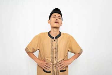 Müslüman kıyafeti giyen Asyalı Müslüman adam acı çekiyor, midesi ağrıyan hareketler yapıyor. İzole edilmiş beyaz arkaplan.