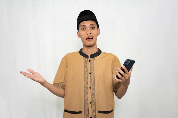 Asian Muslim man wearing Muslim clothes is shocked, confused and surprised while carrying a smartphone. Isolated white background.