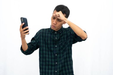 Günlük yeşil kareli gömlek giyen Asyalı bir adam akıllı telefon ekranına bakarken kötü haber aldığı için stresli ya da başı dönüyor. İzole edilmiş beyaz arkaplan.