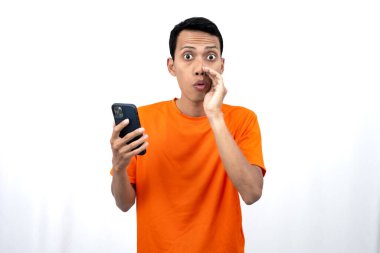 Portakal rengi tişört giyen genç Asyalı bir adamın portresi. Eli ağzının kenarında, dedikodu fısıldıyor ve akıllı telefonu tutarken sırlarını anlatıyor. Haber paylaşımı beyaz arkaplanda izole edildi.