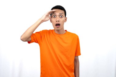 Turuncu tişört giyen genç bir Asyalı 'nın portresi şaşırmış, şok olmuş ve şok olmuş görünüyor. Eller gözlere yakın, beyaz arka planda izole edilmiş..