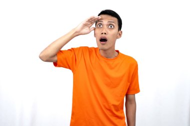 Turuncu tişört giyen genç bir Asyalı 'nın portresi şaşırmış, şok olmuş ve şok olmuş görünüyor. Eller gözlere yakın, beyaz arka planda izole edilmiş..