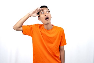 Turuncu tişört giyen genç bir Asyalı 'nın portresi şaşırmış, şok olmuş ve şok olmuş görünüyor. Eller gözlere yakın, beyaz arka planda izole edilmiş..