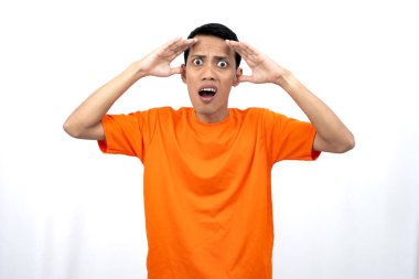 Turuncu tişört giyen genç bir Asyalı 'nın portresi şaşırmış, şok olmuş ve şok olmuş görünüyor. Eller gözlere yakın, beyaz arka planda izole edilmiş..