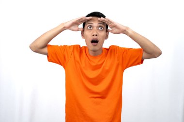 Turuncu tişört giyen genç bir Asyalı 'nın portresi şaşırmış, şok olmuş ve şok olmuş görünüyor. Eller gözlere yakın, beyaz arka planda izole edilmiş..