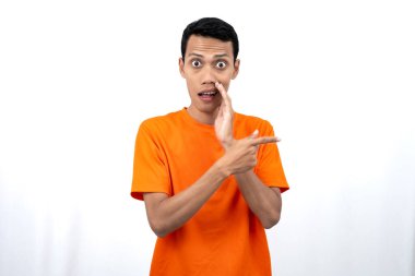Fısıltı pozu vererek fotokopi alanının kenarını gösterirken eli ağzının yanında turuncu tişört giyen Asyalı genç bir adamın portresi. Beyaz arkaplanda izole.