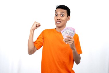 Yüzbinlerce rupiah taşırken mutlu, şaşırmış ve mutlu bir ifadeye sahip turuncu bir tişört giyen Asyalı genç bir adamın portresi. beyaz arkaplanda izole edilmiş.