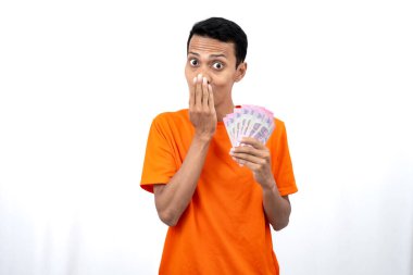 Yüzbinlerce rupiah taşırken mutlu, şaşırmış ve mutlu bir ifadeye sahip turuncu bir tişört giyen Asyalı genç bir adamın portresi. beyaz arkaplanda izole edilmiş.