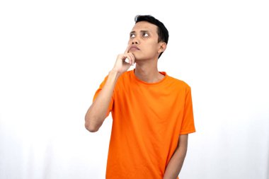 Turuncu tişört giyen, düşünen, fikir arayan ya da kafası karışmış Asyalı genç bir adamın portresi. Beyaz arkaplanda izole.