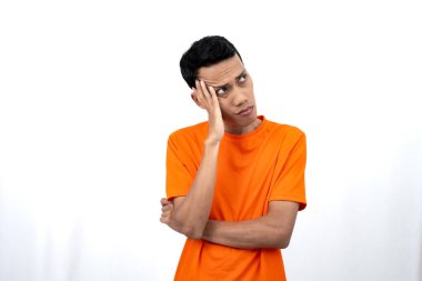 Turuncu tişört giyen, düşünen, fikir arayan ya da kafası karışmış Asyalı genç bir adamın portresi. Beyaz arkaplanda izole.