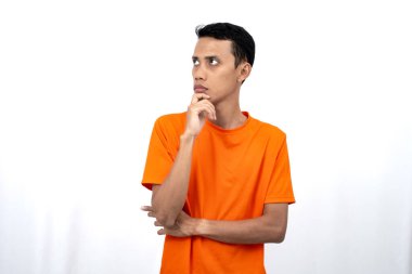 Turuncu tişört giyen, düşünen, fikir arayan ya da kafası karışmış Asyalı genç bir adamın portresi. Beyaz arkaplanda izole.