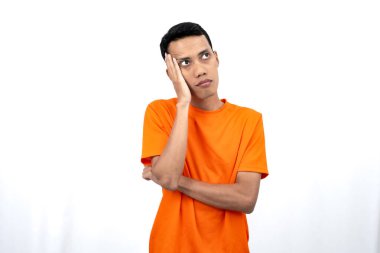 Turuncu tişört giyen, düşünen, fikir arayan ya da kafası karışmış Asyalı genç bir adamın portresi. Beyaz arkaplanda izole.