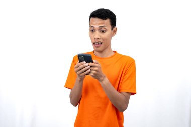 Turuncu bir tişört giyen Asyalı genç bir adamın portresi mutlu bir ifadeyle akıllı telefondan mutlu haberler almasına şaşırdı. Beyaz arkaplanda izole.