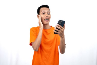Turuncu bir tişört giyen Asyalı genç bir adamın portresi mutlu bir ifadeyle akıllı telefondan mutlu haberler almasına şaşırdı. Beyaz arkaplanda izole.