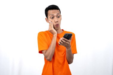 Turuncu bir tişört giyen Asyalı genç bir adamın portresi mutlu bir ifadeyle akıllı telefondan mutlu haberler almasına şaşırdı. Beyaz arkaplanda izole.