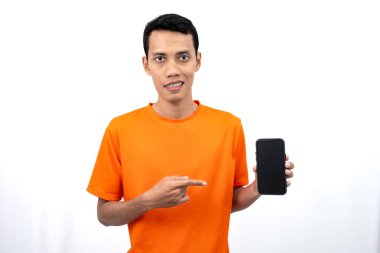 Akıllı telefon ekranını işaret ederken mutlu, şaşkın ve memnun bir ifadeyle turuncu tişört giyen Asyalı genç bir adamın portresi. Beyaz bir arkaplanda izole edilmiş.